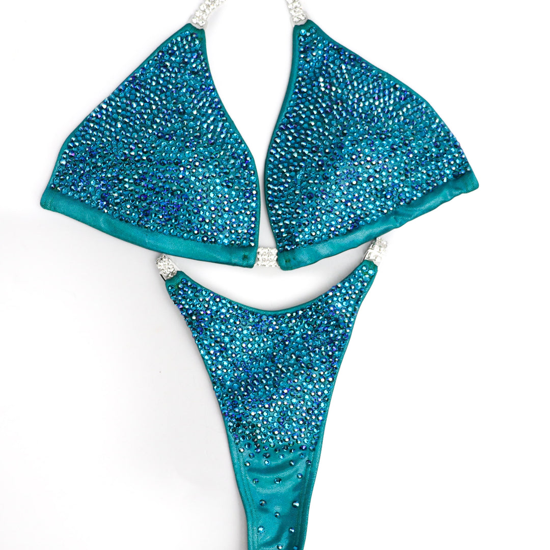 Introducing Casey's Blue Zircon Radiance, a competition suit designed for the fierce female bodybuilder. Inspired by Casey Samsel, its teal and blue hues illuminate the stage with shimmer. Perfect for NPC Bikini or Wellness competitors, this suit exudes confidence and elegance, ensuring you stand out with every pose.