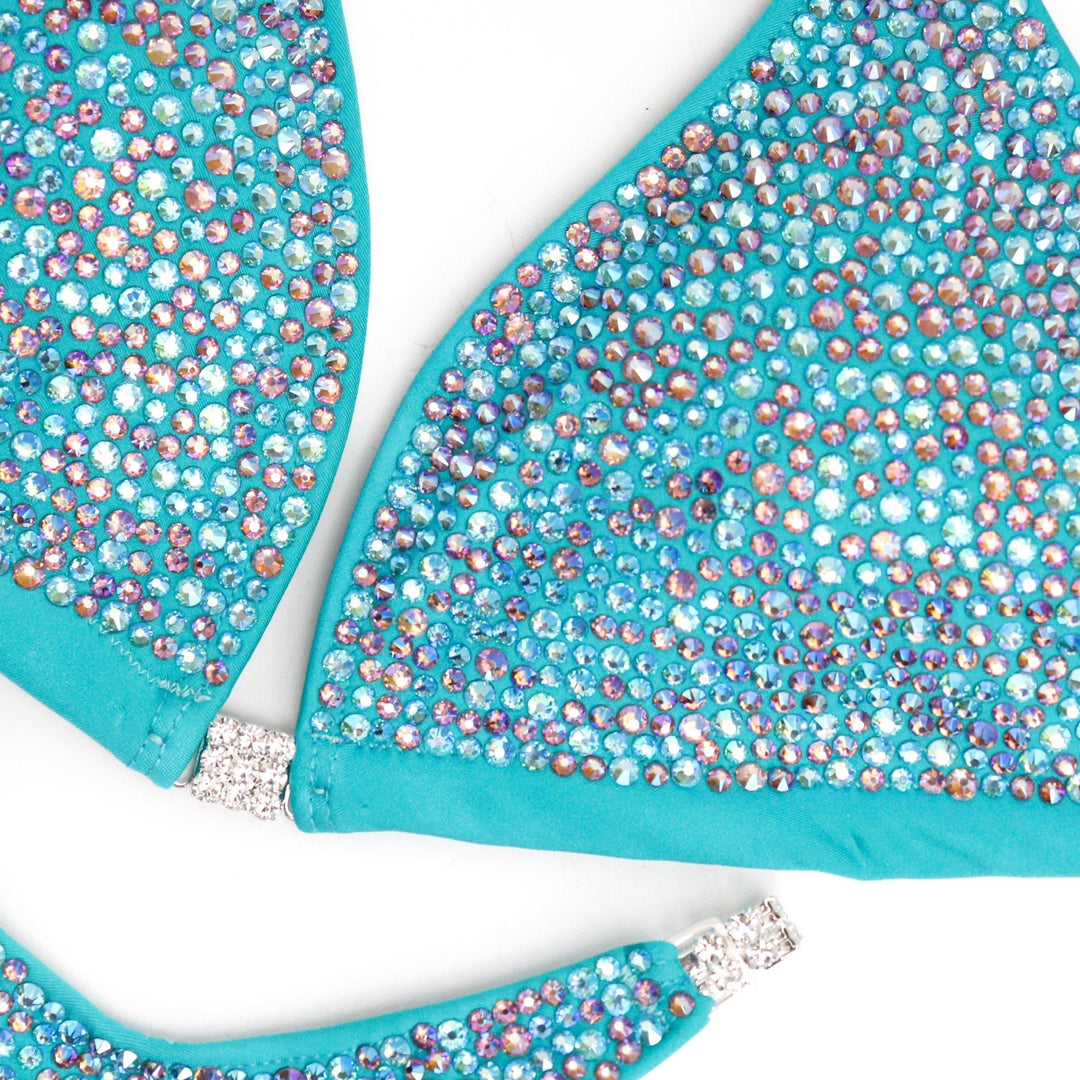 Step into the spotlight with Ashley Hampton's Aqua Radiance! Tailored for NPC Bikini or Wellness competitors, this suit dazzles with bright aqua tones and delicate hints of light amethyst ab. Elevate your stage presence with its radiant design, ensuring you shine like never before.