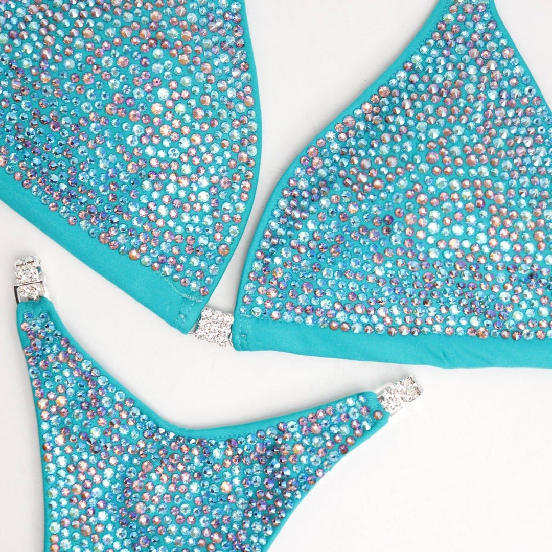 Step into the spotlight with Ashley Hampton's Aqua Radiance! Tailored for NPC Bikini or Wellness competitors, this suit dazzles with bright aqua tones and delicate hints of light amethyst ab. Elevate your stage presence with its radiant design, ensuring you shine like never before.